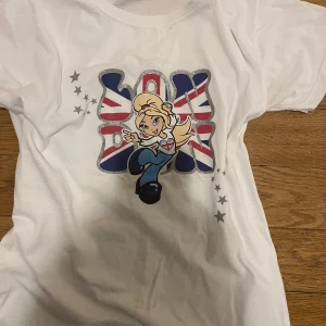 Vit t-shirt med tryck - Säljer en vit t-shirt med ett färgglatt tryck av en tecknad figur och text i brittiska flaggans färger. T-shirten har korta ärmar och är perfekt för en avslappnad stil. Stjärnor i silver pryder sidan av trycket.