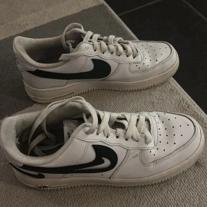 Vita Nike Air Force sneakers - Säljer ett par klassiska vita Nike Air Force sneakers med svart swoosh. Skorna har snörning och perforeringar på tån för ventilation. Perfekta för en stilren look.