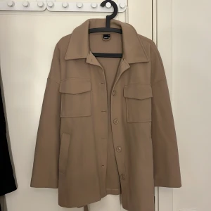 Beige kappa med bälte - Snygg beige kappa med knappar och bälte i midjan. Jackan har långa ärmar och stora fickor framtill. Perfekt för en stilren look. Storlek M och har används 3-5 gånger.