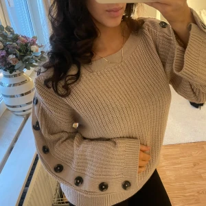 Beige stickad tröja med knappar - Säljer en mysig beige stickad tröja med stora svarta knappar längs ärmarna. Tröjan har en avslappnad passform och långa ärmar, perfekt för kyligare dagar. En stilren och bekväm tröja som passar till många tillfällen.