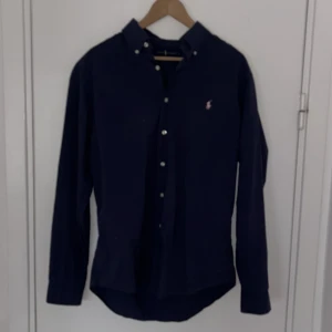 Mörkblå skjorta från Ralph Lauren - Snygg mörkblå skjorta från Ralph Lauren i slim fit. Tillverkad i bomull med stretch för extra komfort. Skjortan har långa ärmar och knappar framtill. Den ikoniska broderade loggan i rosa ger en stilren touch.