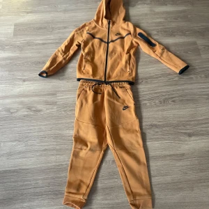 Orange hoodie från Nike Tech - Säljer en snygg orange hoodie och byxor från Nike med dragkedja och huva. Perfekt för träning eller en avslappnad dag. Den har svarta detaljer och en ficka på ärmen. Barnstorlek S (128-137)