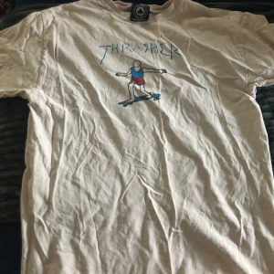 Vit t-shirt från Thrasher - Säljer en lite lätt rosa t-shirt från Thrasher med ett coolt tryck av en tecknad figur på skateboard. T-shirten har korta ärmar och är perfekt för en avslappnad stil. Passar bra för skate-entusiaster! Den har blivit lite missfärgad i tvätten så den har en ljusrosa färg numera