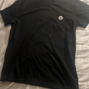 Svart t-shirt från Moncler - Säljer en stilren svart t-shirt från Moncler med en liten logotyp på bröstet. Perfekt för en enkel och snygg look. T-shirten har korta ärmar och en rund halsringning.