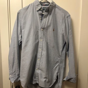 Blå skjorta från Polo Ralph Lauren - Snygg blå skjorta från Polo Ralph Lauren med klassisk krage och knappar framtill. Den har en broderad logga på bröstet och långa ärmar. Perfekt för en stilren look.