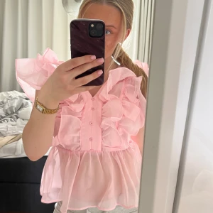 Rosa volangblus med knappar - Supersöt rosa blus med volanger och knappar framtill. Blusen har korta puffärmar och är tillverkad i ett tunt, lätt material som ger en luftig känsla. 