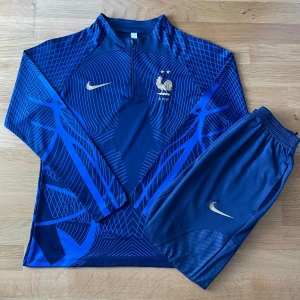 Frankrike fotbollströja och byxor  Nike - Mörkblå långärmad Frankrike fotbollströja med matchande byxor. Tröjan har blått mönster, guldiga Nike-loggor och FFF-emblem med tupp och två stjärnor. Materialet är syntetiskt och snabbtorkande, perfekt för träning.