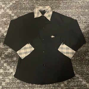 Svart skjorta från Burberry -  svart skjorta från Burberry med klassiskt rutigt mönster på krage och ärmslut. Den har ett litet hål på sidan men syns knappt för att jag har sytt. Storlek M i barns storlek! Vid frågor är det bara ställa svara på alla🤩