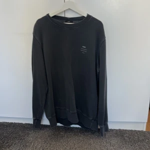 Mörk Grå sweatshirt från Neuw - Mörk Grå sweatshirt från Neuw med rund halsringning och diskret tryck på bröstet. Tröjan har långa ärmar och klassisk passform, perfekt för en avslappnad stil. Mjuk bomullskänsla och ribbade muddar vid ärmslut och nederkant.