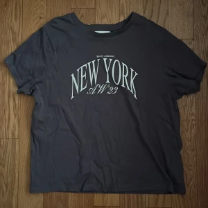 Mörkgrå t-shirt med New York-tryck från Pull&Bear - Säljer en mörkgrå t-shirt från Pull&Bear med stort vitt New York-tryck på bröstet. Klassisk passform med rund hals och korta ärmar. Perfekt för en chill och stilren look.