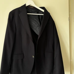 Mörkblå blazer från Oscar Jacobson - Klassisk mörkblå blazerfrån Oscar Jacobson med två fickor framtill och en knappstängning. Perfekt för formella tillfällen och stilrena outfits. Tunn fodring och diskret struktur i tyget. Finns hål för att sy dit en till knapp som ligger i ena fickan 