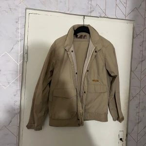 Beige vindjacka med dragkedja - Säljer en beige vindjacka med klassisk krage och dragkedja framtill. Jackan har två stora fickor framtill och ribbad nederkant. Perfekt för dig som vill ha en stilren och enkel jacka till vardags.