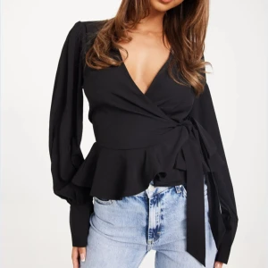 Svart omlottblus med knytband - Snygg svart omlottblus med v-ringning och långa ärmar. Blusen har ett brett knytband i midjan och volangdetalj nedtill. Perfekt att styla med jeans eller kjol.
