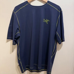 Blå funktions-t-shirt från Arc'teryx med grafiskt tryck - Säljer en blå t-shirt från Arc'teryx i storlek L. T-shirten har gula kontrastsömmar, liten logga fram och stort grafiskt tryck i gult på ryggen. Perfekt för dig som gillar snygga detaljer och sportig stil.