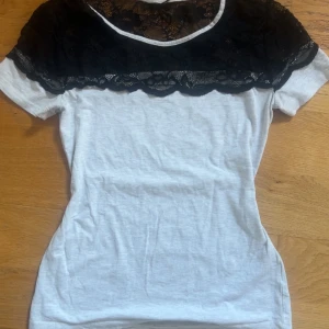 Ljusgrå t-shirt med svart spets - Säljer en ljusgrå t-shirt med svart spets upptill och över axlarna. T-shirten har korta ärmar och en rund halsringning. Perfekt för dig som vill ha en enkel topp med en snygg detalj. Den är mest intil vit som man ser på bild.