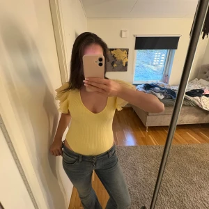 Body Zara - Säljer en ljusgul ribbad topp från Zara med djup v-ringning och volangdetaljer på ärmarna. Toppen är tight och har korta ärmar med snygga volanger som ger en extra touch. Perfekt för dig som vill sticka ut lite extra! Perfekt till sommaren 