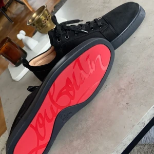 Svarta sneakers  - Säljer ett par svarta sneakers från Christian Louboutin med klassisk röd sula och rund tå. Skorna har snörning och är tillverkade i mocka med svarta detaljer. Perfekta för dig som vill sticka ut med en lyxig touch.