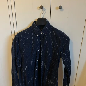 Gant skjorta - Snygg mörkblå skjorta från GANT med små vita kryssmönster. Skjortan har klassisk krage med knappar och lång ärm. Perfekt för dig som gillar stilrena detaljer och vill ha något som sticker ut lite.