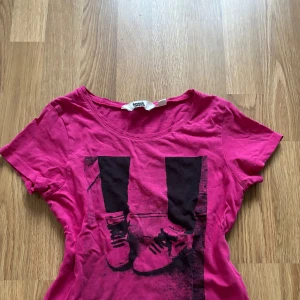 Rosa t-shirt med tryck från Heart Music - Cool rosa t-shirt från Heart Music med svart tryck av ben sneakers på framsidan. T-shirten har rund halsringning och är kortärmad. Perfekt för dig som gillar streetstyle och vill sticka ut med en färgstark look. 