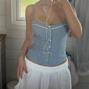 Blåvit rutig korsettopp - Säljer en blåvit korsettopp med rutigt mönster och smala axelband. Toppen har knappar framtill och en figurnära passform som framhäver midjan. Perfekt för sommardagar och att styla med kjol eller jeans.