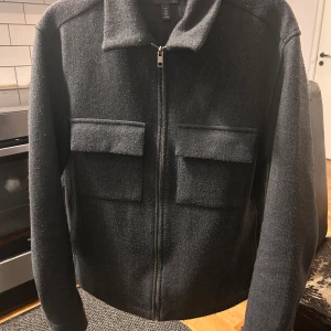 Svart jacka med dragkedja från H&M - Stilren svart jacka med klassisk krage och två stora bröstfickor. Jackan har dragkedja framtill och är tillverkad i ett mjukt material med ullkänsla. Perfekt för kyligare dagar och enkel att matcha med olika outfits. Kan sänka vid snabb affär