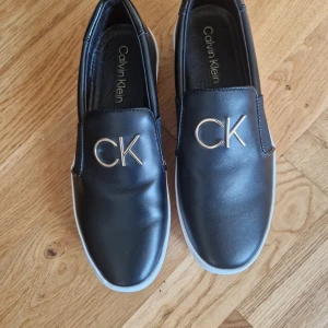 Svarta slip-on sneakers från Calvin Klein - Snygga svarta slip-on sneakers från Calvin Klein med vita platta sulor och guldfärgade CK-detaljer på ovansidan. Skorna har en stilren design i syntetmaterial och är perfekta för en trendig vardagslook.