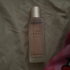 La Rive I Am Ideal Eau de Parfum - La Rive I Am Ideal är en eau de parfum för kvinnor. Flaskan är rektangulär med en transparent design och ett silvrigt räfflat lock. Doften är elegant och flaskan har en stilren, modern look med text i svart och guld.