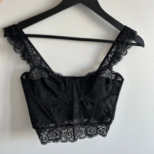 Svart spetsig korsett-topp - Säljer en svart korsett-topp i spets med axelband och spetsdetaljer. Toppen har markerade kupor och vacker kant nertill. Går att bära både med eller utan bh. Perfekt för att lyfta en outfit. Bara hängt i garderoben så skicket är som ny!❤️