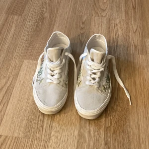 Beiga sneakers från Vans med broderade blommor - Säljer ett par beiga Vans sneakers med broderade blommor i grönt och beige på sidorna. Skorna har snörning och platt sula. Perfekta för dig som gillar unika detaljer! Nypris: 849:- 