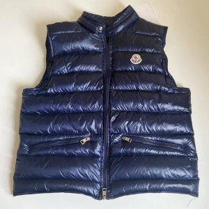 Mörkblå dunväst från Moncler - Snygg mörkblå dunväst från Moncler. Västen har två fickor med dragkedja och Moncler-logga på bröstet. Perfekt för lager-på-lager och stilren look. Dessvärre saknas 1 utav de 2 dragkedjorna som ska finnas på västen, borde gå att fixa om man pratar med någon som vet hur man fixar det. 