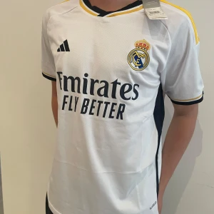 Real Madrid fotbollströja 2023/24 - Säljer en vit Real Madrid hemmatröja från Adidas med svarta och gula detaljer. Tröjan har klubbmärke och sponsortryck på bröstet samt korta ärmar. Klassisk design med gula ränder på axlarna och svart krage.