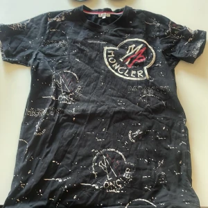 Svart t-shirt från Moncler - Svart t-shirt från Moncler med stort broderat logotyptryck på bröstet och mindre tryck över hela tröjan.