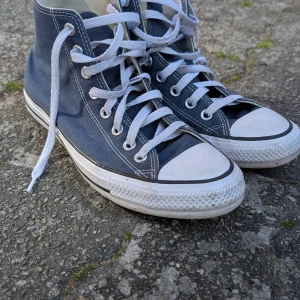 Mörkblå Converse Chuck Taylor All Star - Säljer ett par klassiska mörkblå Converse Chuck Taylor All Star med högt skaft och vita snören. Skorna har vit gummisula, rund tå och den ikoniska loggan på sidan. Perfekta för dig som gillar tidlös streetstil.