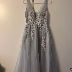 Ljusgrå balklänning med spetsdetaljer - Elegant ljusgrå balklänning med djup v-ringning och vackra broderade spetsdetaljer. 