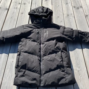 Svart dunjacka från Sail Racing - Säljer en svart dunjacka från Sail Racing med huva och dragkedja. Jackan har en bröstficka med dragkedja och Sail Racing-logga samt patch med 'Sail Antarctica'. Perfekt för kalla dagar och riktigt varm tack vare dunfyllning.