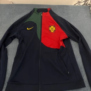 Portugal Nike fotbollströja med dragkedja - Säljer en Nike Portugal fotbollströja med dragkedja. Tröjan är långärmad och har färgblock i mörkblått, rött och grönt med Portugals landslagsemblem på bröstet. Slim fit-modell med gul Nike-logga. Kan diskutera pris! (Äkta)