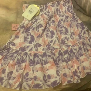 Blommig volangkjol från By Malina - Supersöt kort kjol från By Malina med blommigt mönster i lila och rosa toner. Kjolen har bred resår i midjan och volang nertill som ger en luftig känsla. Perfekt för varma dagar och enkel att matcha med en enkel topp.