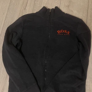 Marinblå zip-jacka från Hugo Boss - Snygg mörkblå zip-jacka från Hugo Boss med röd logga på bröstet. Jackan har hög krage, dragkedja framtill och ribbade muddar. Perfekt för en stilren och avslappnad look. Pris kan diskuteras!