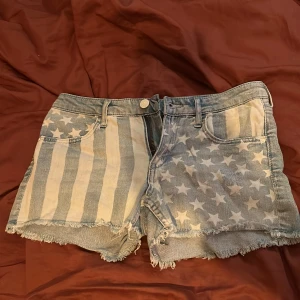 Jeansshorts med amerikanskt flaggmönster - Säljer ett par coola jeansshorts med fransig kant. Ena sidan har vita stjärnor på blå botten och andra sidan har vita och blå ränder, inspirerat av amerikanska flaggan. Perfekta för sommaren och festivaler. 74 cm i midja och lågmidjade