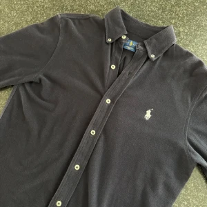 Mörkblå skjorta från Ralph Lauren - Klassisk mörkblå skjorta från Ralph Lauren med knappar hela vägen och broderad logga på. Tillverkad i meshmaterial. Perfekt för en stilren look till alla årstider. Köpt på Johnells för 1800kr