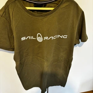 Olivgrön t-shirt från Sail Racing - Olivgrön t-shirt från Sail Racing med vit logotyptryck på bröstet. Klassisk rund halsringning och korta ärmar. Perfekt för en avslappnad och sportig stil.