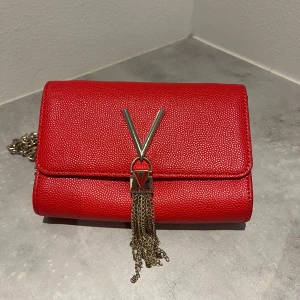 Röd axelväska från Valentino - Säljer en snygg röd axelväska från Valentino med guldfärgad kedja och dekorativt V-spänne framtill med hängande kedjor. Väskan har en stilren och kompakt design, perfekt för att lyfta din outfit. 