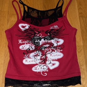 Y2K Vinröd linnetopp med spets och tryck - (Meddela innan köp) Säljer en vinröd linnetopp med svarta spetsdetaljer och tryck framtill med vingar, hjärta och texten 'Love'. Toppen har smala axelband och spetskant nertill, det svarta spets tyget som är under sitter ihop med det röda, säljer eftersom att jag tycker att den sitter för tajt på mig❤️