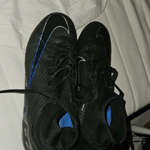 Svarta fotbollsskor Nike Mercurial - Säljer ett par svarta Nike Mercurial fotbollsskor med blå och vita detaljer. Skorna har snörning och en smal passform med rund tå. Perfekta för dig som vill ha lätta och snabba skor på planen.