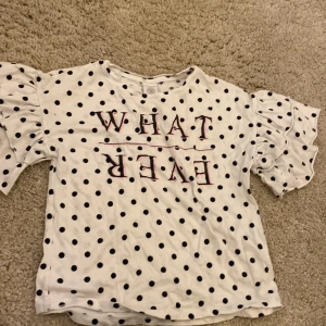 Vit prickig t-shirt med puffärmar från Lindex - Säljer en vit t-shirt från Lindex med svarta prickar och texten 'WHATEVER' i rosa och svart. Tröjan har korta puffärmar som ger en trendig look. Perfekt för dig som gillar statement-plagg och mönster.