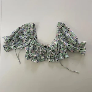 Blommig croppad blus med volang - Superfin blommig croppad blus med volangdetaljer och puffiga trekvartsärmar. Blusen har knytband framtill och ett romantiskt mönster i pastellfärger. Perfekt för vår och sommar!