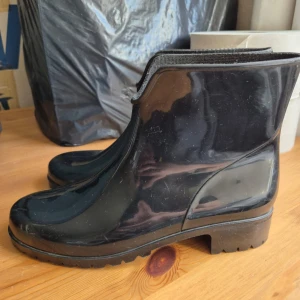 Svarta gummistövletter - Säljer ett par svarta, blanka stövletter i gummi. Skorna har rund tå, platt sula och är vattentäta – perfekta för regniga dagar. De är enkla att dra på och av tack vare den korta skaftmodellen.