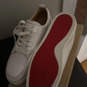Vita sneakers med röd sula från Christian Louboutin - Säljer ett par stilrena vita sneakers från Christian Louboutin med den ikoniska röda sulan. Skorna har rund tå, vita skosnören och är tillverkade i skinn. Perfekta för dig som vill ha en klassisk men lyxig look.