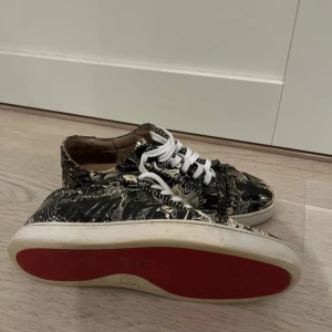 Svartvita sneakers med grafiskt mönster och röd sula - Säljer ett par unika sneakers från Christian Louboutin med svartvitt grafiskt mönster och klassisk röd sula. Skorna har vita snören och rund tå. Perfekta för dig som vill sticka ut med din stil.