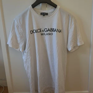 Vit t-shirt från Dolce & Gabbana - Säljer en klassisk vit t-shirt från Dolce & Gabbana med svart logotyptryck på bröstet. T-shirten har rund halsringning och korta ärmar. Perfekt för en stilren och enkel look.